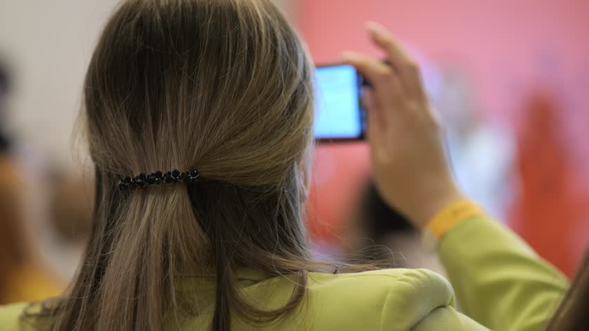 A woman with long brown hair is taking a picture with her cell phone. She is wearing a green jacket and is standing in front of a red wall