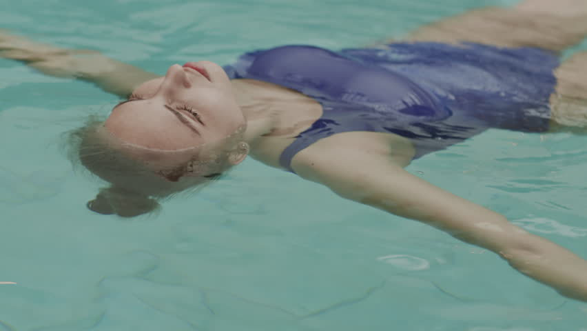 Lockdown of young Caucasian woman wearing blue swimming suit lying on her back on water surface in indoor pool, looking up and relaxing
