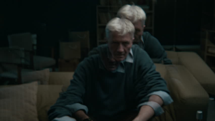 Low key shot of elderly Caucasian man sitting in dark living room covering his face with hands having anxiety attack at home and feeling overwhelmed