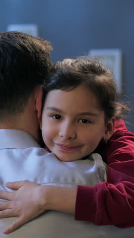 Adult man hugging little girl looking into camera. Loving father and kid girl embrace, bonding and smile together on the sofa at home. Vertical shot