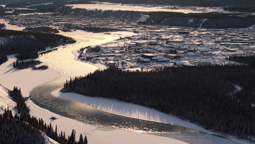 A smooth aerial tilt-up establishing shot at sunset, capturing Downtown Whitehorse, the winding Yukon River, and the rugged, untouched landscape surrounding the city in this peaceful northern setting.