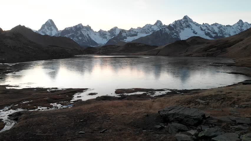 Aerial flyover over a half frozen lake during an autumn sunset with a reflection of mountains at Lacs de Fenêtre in Valais, Switzerland with a view of snow-capped peaks of the Alps such as Mont Blanc.