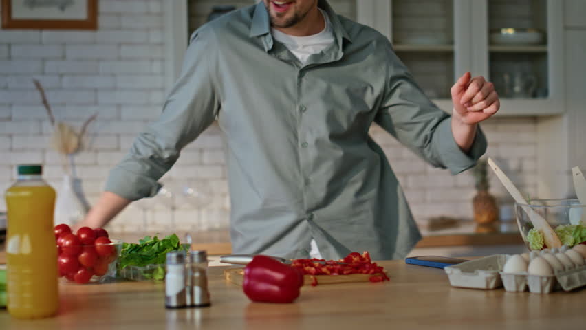 Guy preparing food in headphones dancing along favorite song at cozy kitchen closeup. Carefree joyful man listening music in headset cooking breakfast. Smiling brunette cutting vegetables at home