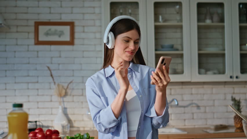 Lady using smartphone kitchen reading message while cooking closeup. Joyful woman in headphones listening music turning song at cellphone. Smiling girl in earphones enjoying culinary with mobile phone