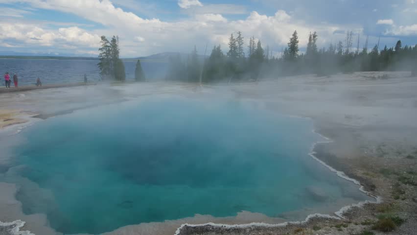 View of a bright turquoise hot spring at the West Thumb Geyser Basin, with steam emanating from its surface while people walk on a boardwalk.