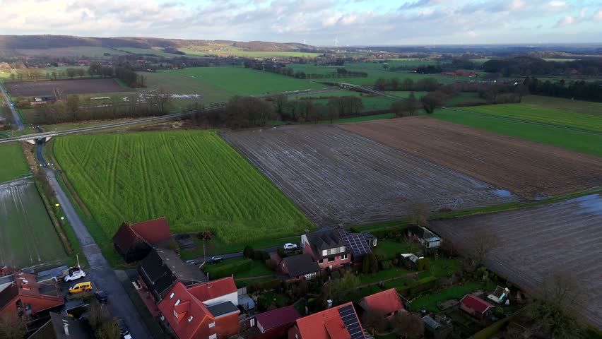 Aerial shot of houses and homes in rural area of USA. Solar panels on roof. Wet agricultural farm fields and highway in distance. Daytime with clouds in winter. Wide shot.