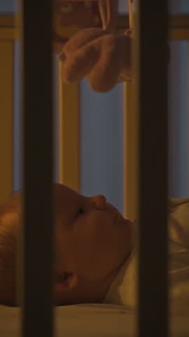 A baby lying in a crib gazes up in wonder at a soft toy held above. The warm, gentle lighting of the nursery creates a peaceful atmosphere, perfect for bedtime exploration