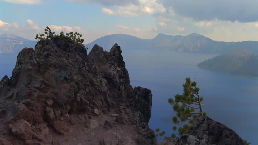 Rocky cliff with small pine trees overlooking Crater Lake’s deep blue waters. Oregon, USA volcanic lake and famous tourist location