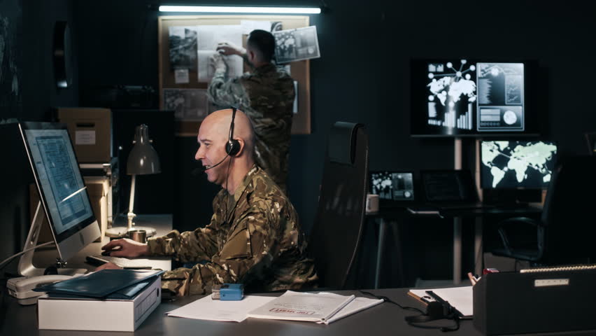Pan shot of adult military agent in headset transmitting information in front of computer screen with blueprint while his colleague working with noticeboard in command center