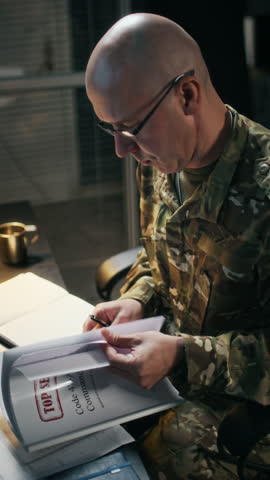 Vertical shot of adult Caucasian commander in camouflage uniform working with top secret documents at command hub
