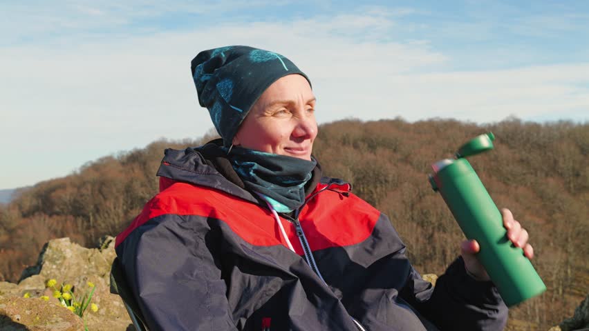 Girl tourist resting on a rocky ledge, dressed in a brightly colored jacket and hat. In her hand she has a green thermos with tea from which she takes a sip. In the background are picturesque hills