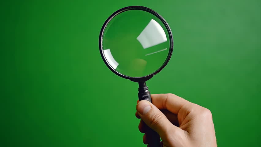 Hand holds classic magnifying glass against green chroma key backdrop. Lens loupe reflects light, focus point. Magnifier exploration, curiosity. Investigation, research, education. Discovery learning.