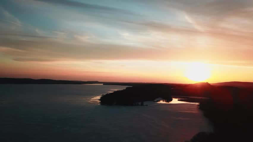 A stunning winter sunset casting warm hues over a frozen lake surrounded by silhouettes of trees and hills.