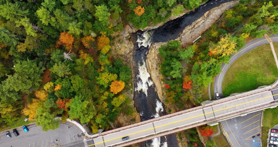 Descending above the bridge over the narrow river with rapids. Beautiful colorful trees grow around the river.