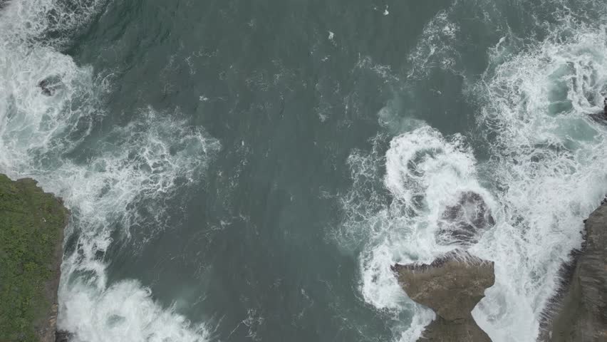 Aerial view of rocky shoreline with a large wave crashing into the rocks. The water is white and the rocks are grey