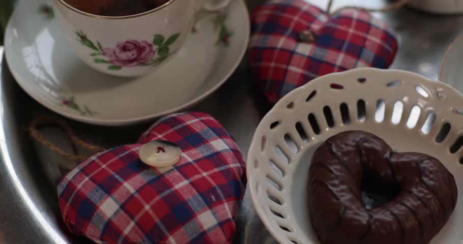 Heart shaped chocolate dessert on plate closeup. Cup of tea and cookie on tray. Hand takes biscuit and put with bite. Handcrafted fabric stuffed hearts. Cooked dessert with beautiful presentation.
