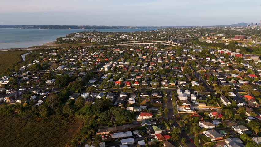 Houses and homes in suburb district of Auckland city during sunset time. Cozy ambiance and atmosphere in New Zealand. Bridge and water in distance. Aerial forward wide shot.