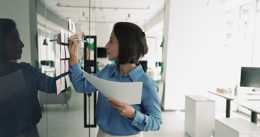 Focused woman office employee holds paper, review written information, make notes on sticky notes attached to wall, structuring to-do list, setting goals, busy in project management, task organization
