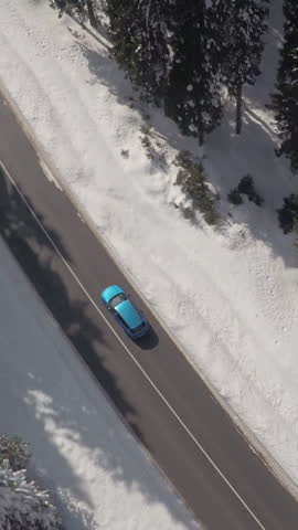 AERIAL: Flying above turquoise sports car driving along narrow highway in deep snowy mountain forest in wintertime. Road trip through pine tree woodland covered with white snow on sunny Christmas day