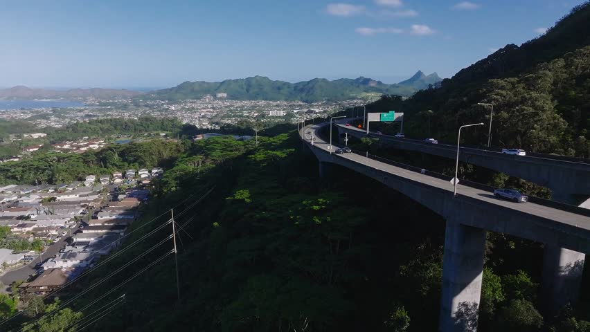 Aerial view of a state highway curving through green hills on Oahu, Hawaii, with moving vehicles, Honolulu cityscape, and Ko olau mountains under a clear sky.