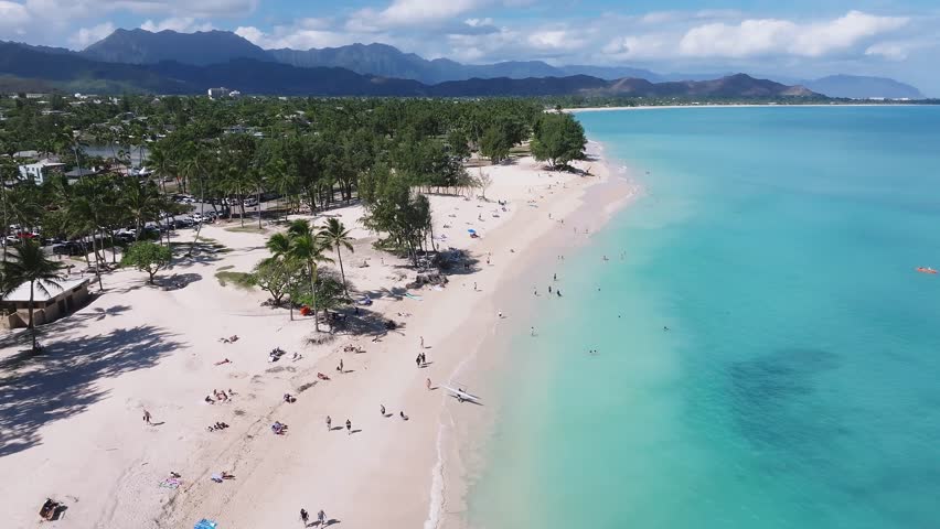 Aerial view of a Kailua beach on Oahu, Hawaii, featuring turquoise waters, beachgoers, a paddleboarder, lush greenery, and the Ko olau mountain range.