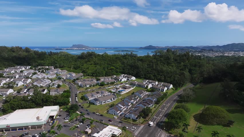 Aerial view of a residential area on Oahu island, Hawaii, with houses, lush greenery, turquoise ocean, distant mountains, and a road with light traffic.
