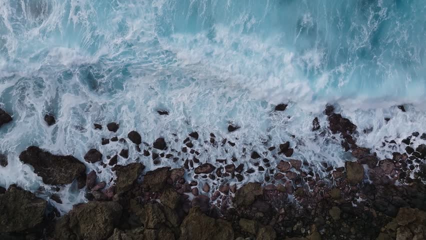 Dynamic aerial view of waves breaking against rugged rocks on Oahu Island, Hawaii, showcasing the contrast of ocean blues and earthy tones.