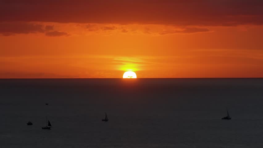Sunset over the ocean near Oahu, Hawaii, with vibrant orange and red skies. Sailboats and small vessels gently move across the calm water.