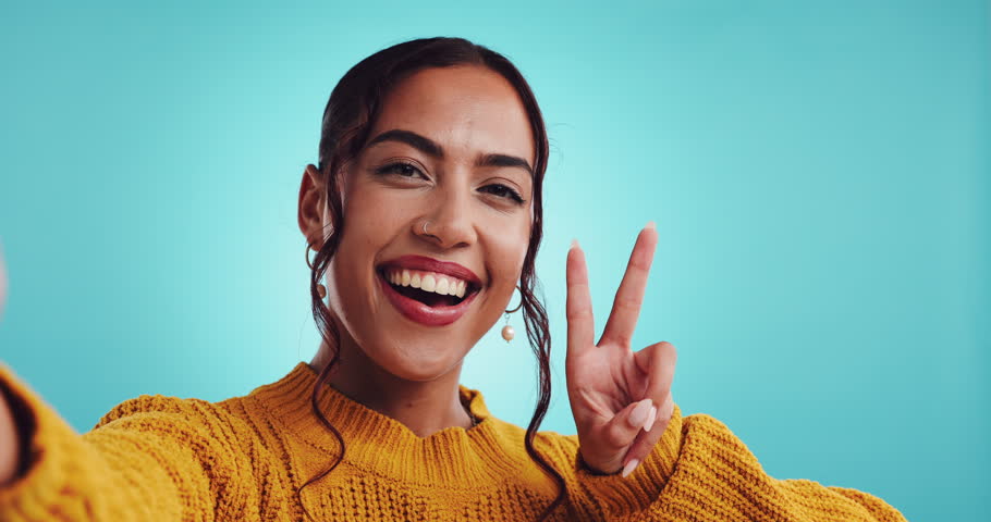 Happy woman, peace sign and face in studio for selfie, video call and social media update. Influencer, hand V and confident on blue background for photography, connectivity and dating app profile