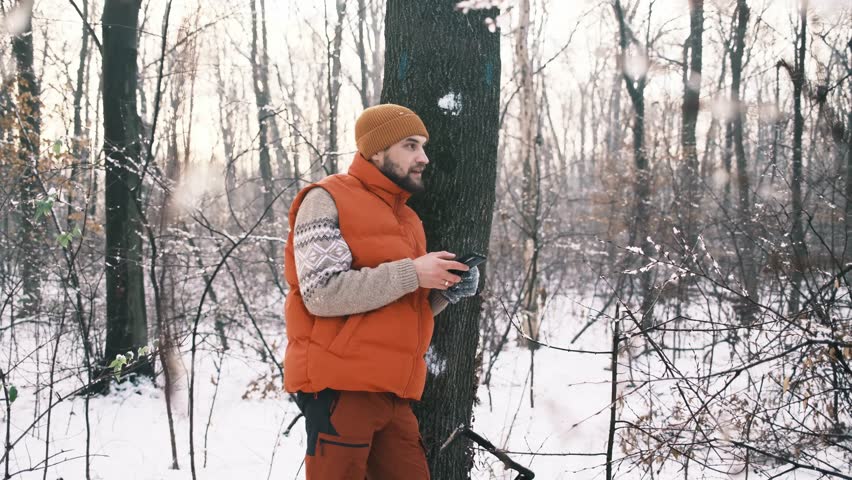 A young Caucasian man holds a phone in his hands and watches a video while leaning on a tree in a snowy winter forest. Wireless technology concept
