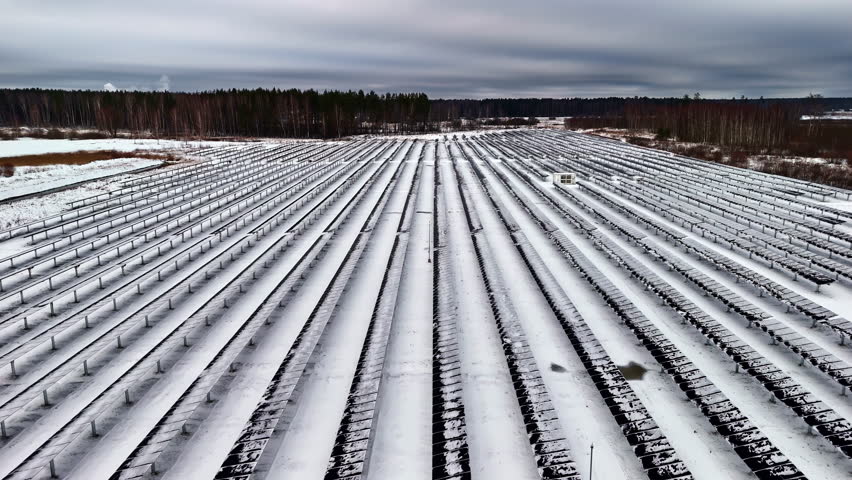 Snow capped rows of solar panel, solar park for renewable green energy, future power in winter, sun, dolly shot forward above the panels, grey cloudy sky