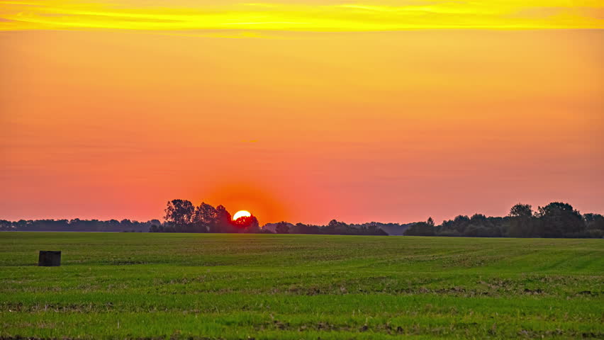 Sunrise timelapse over a grass field. Bright, orange sunlight shines on an open rural landscape.