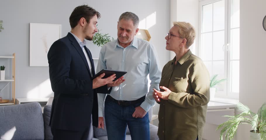 Young male real estate agent giving a tour about the house or apartment to a nice retired senior family couple investors. Real estate business concept 