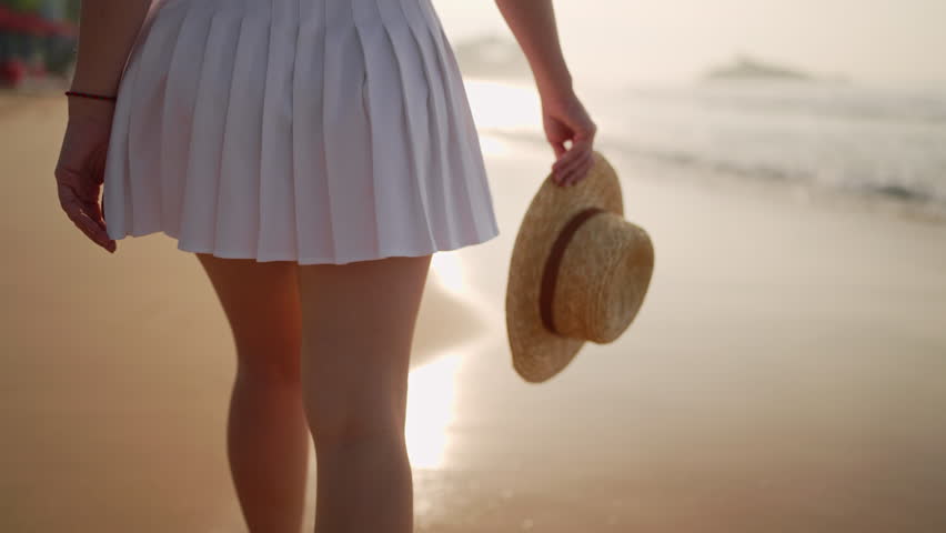 Woman holds straw hat walking on sandy beach at sunset. Female in white skirt enjoys seaside stroll, waves gently wash ashore. Leisure travel, serene sea scene.