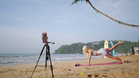 Fitness instructor conducts beach yoga class via livestream. Blonde in sportswear demonstrates plank on mat, smartphone on tripod records. Interactive home training, personalized exercise guidance. - Powered by Shutterstock - Get 15% off with code: PIKWIZARD15