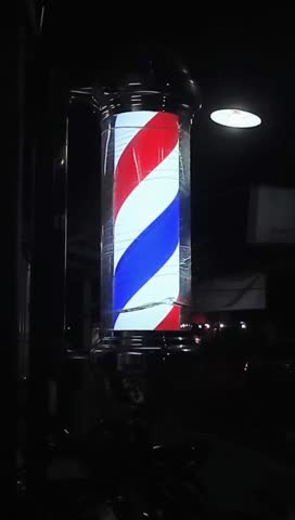classic barber pole bathed in the darkness of night, illuminated by a soft light source. The red, white, and blue stripes spiral upwards, symbolizing the traditional trade of barbering.