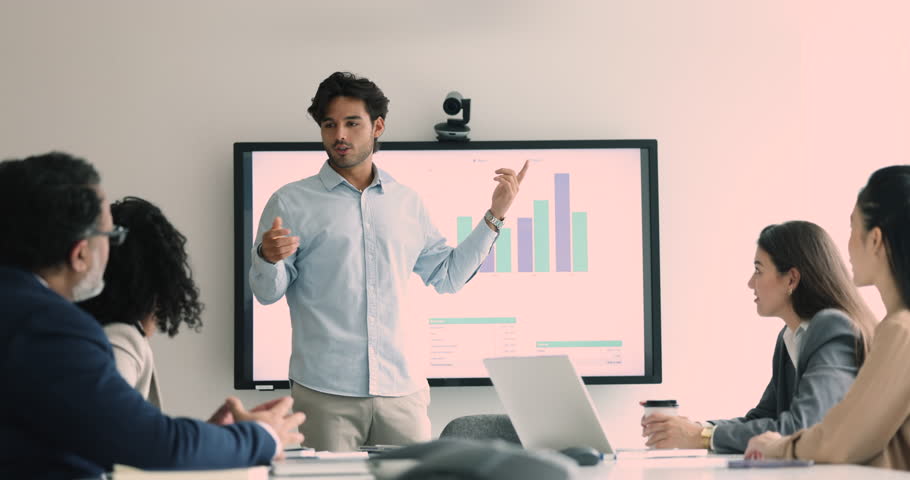 Confident young Latin project manager man presenting sales growth reports, showing presentation business partners, investors, standing at electronic board, talking to diverse business colleagues