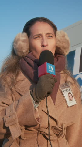 Medium vertical shot of Caucasian female journalist in earmuff headphones, winter coat, talking into microphone while delivering broadcast news report for local TV channel