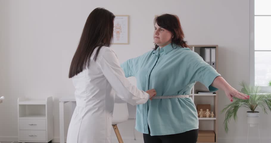 Young doctor nutritionist measure waist with measuring tape for a fat plump overweight woman patient standing in medical clinic during examination. Slimming, weight loss and diet concept. 4k video.