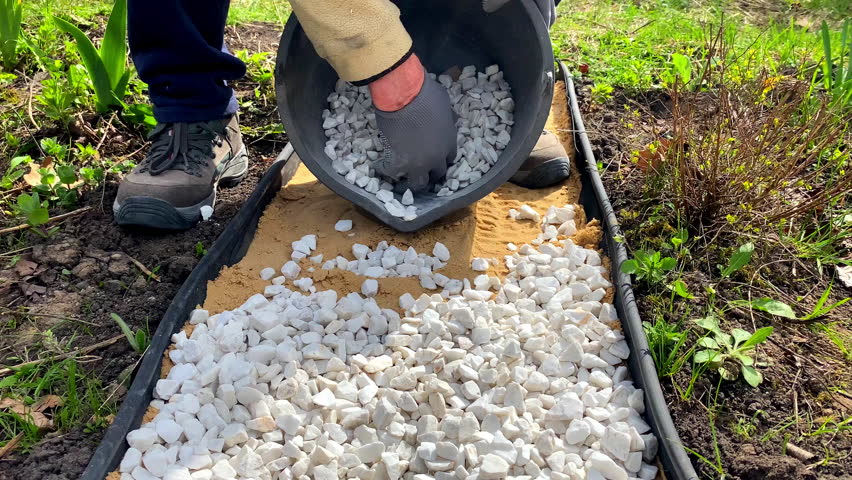 Man worker strewing white decorative split gravel, stones,rocks on path,road,walkway,pathway made of sand on flower bed lawn in garden, landscape design.