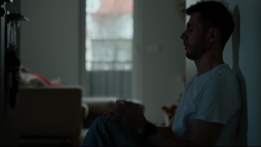 Depressed young man sitting on floor in dark room. Silhouette of young male suffering from loneliness and anxiety, covering his face with hands. Person with mental disorder