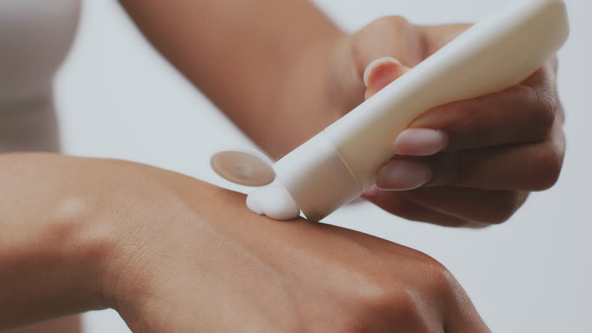 A millennial African American woman is pampering herself at home, applying a skin treatment to her arm. She focuses on self-care and enhancing her natural beauty in a serene environment.