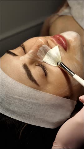 A beautician applies a white cleansing peeling mask to a female face close-up, vertically. A woman at a relaxing spa treatment enjoys