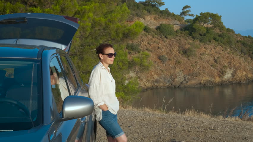 Admiring beautiful seascape by car. A happy lady with pleasure stand by the car and look around the sea nature from the bay.
