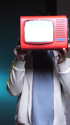 vertical video man has a TV instead of a head. Man is holding a small Red portable television in front of his face. Obscuring his identity against a dimly lit. Teal backdrop. 