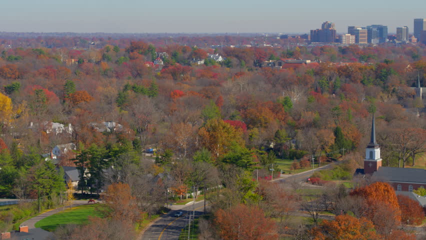 Aerial view over Clayton Rd in St. Louis, Missouri, featuring a charming church surrounded by vibrant autumn trees. The stunning fall colors frame the downtown Clayton skyline on the horizon.