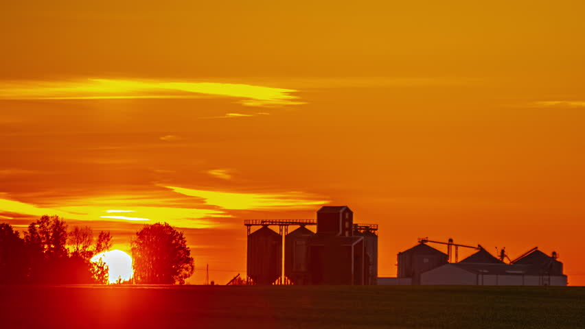Timelapse of orange sunrise behind a silhouette of trees and grain silos on farm