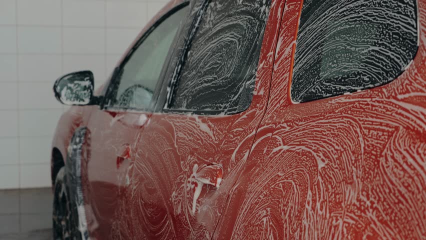 Red car getting a foamy wash at a self-service car wash. Soap suds cover the vehicle as it gets cleaned. Car wash, soap, water, clean, vehicle.