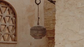 An ornately designed metal lantern hangs from a wrought iron hook outside a building. Beige walls and lattice window create a tranquil Moroccan scene. This traditional decor adds a touch elegance - Powered by Shutterstock - Get 15% off with code: PIKWIZARD15