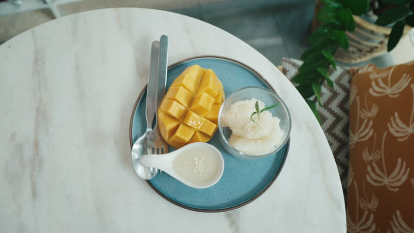Mango sticky rice is elegantly presented on a blue plate, accompanied by a small bowl of coconut cream. Warm and inviting atmosphere, dessert lovers. Thai food. Top view